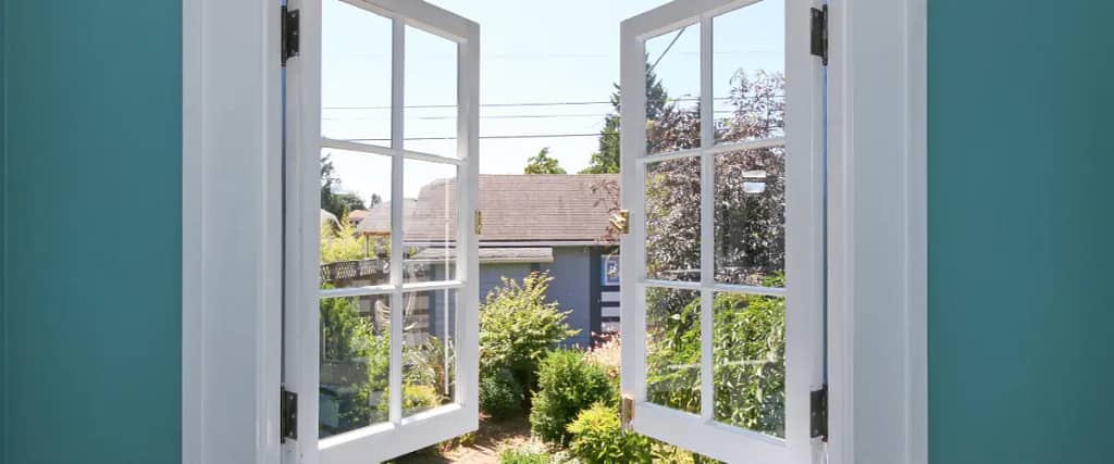 Open white-framed windows overlooking a garden with shrubs and a neighboring house in the background.